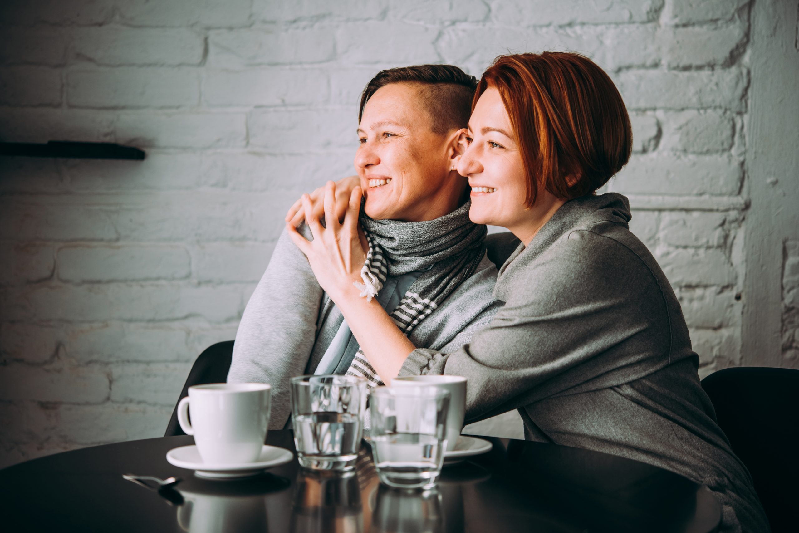 shutterstock_1055875097 Photo of two women sitting at a table having a coffee. Both women are looking towards the same direction, one woman is hugging the other. Both women are smiling.