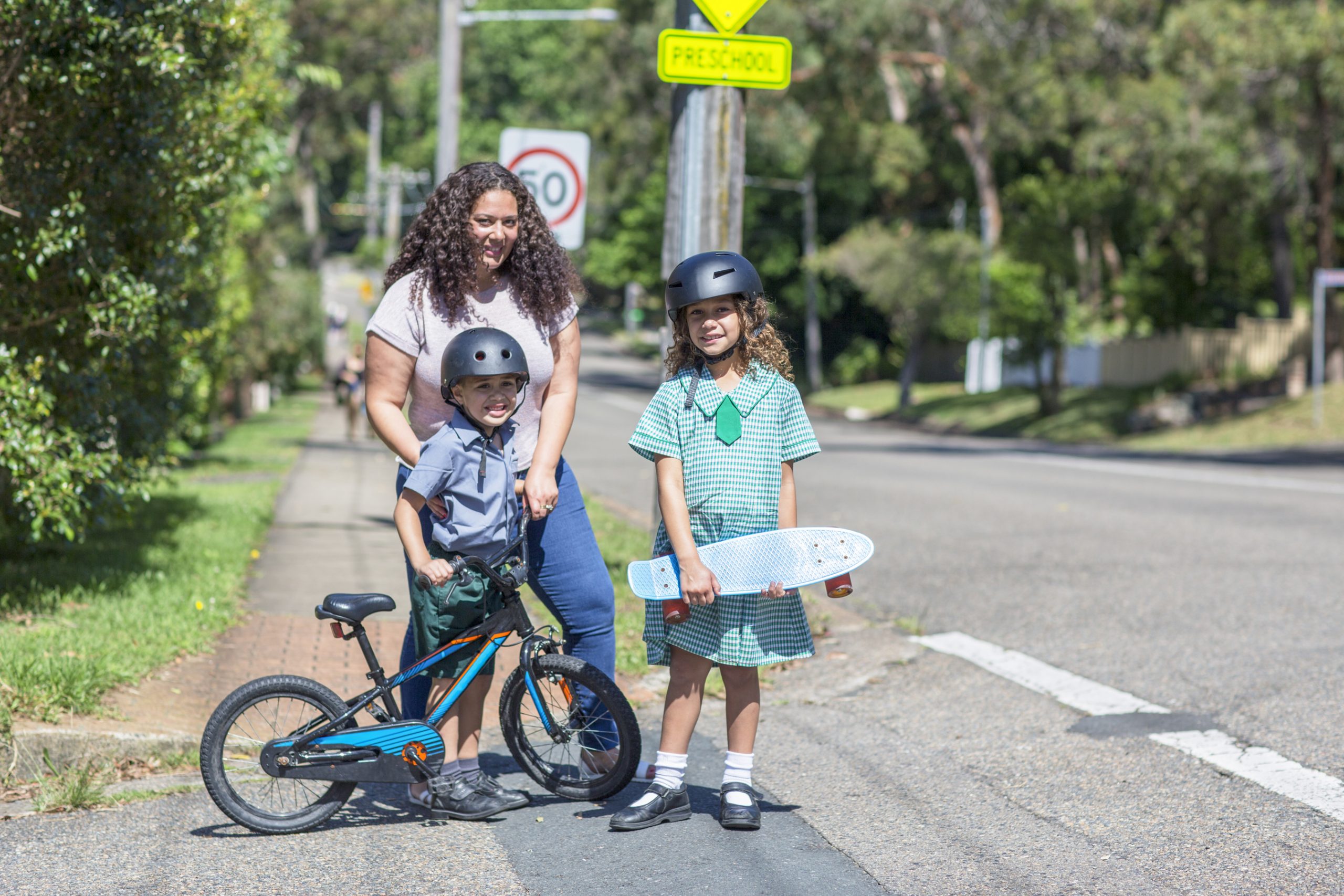Woman with long dark hair standing on a road, with two children - a young boy aged about 6 years old on push bike and wearing a helmut, a young girl aged 11 years old wearing her school uniform, a helmut and carrying a skateboard