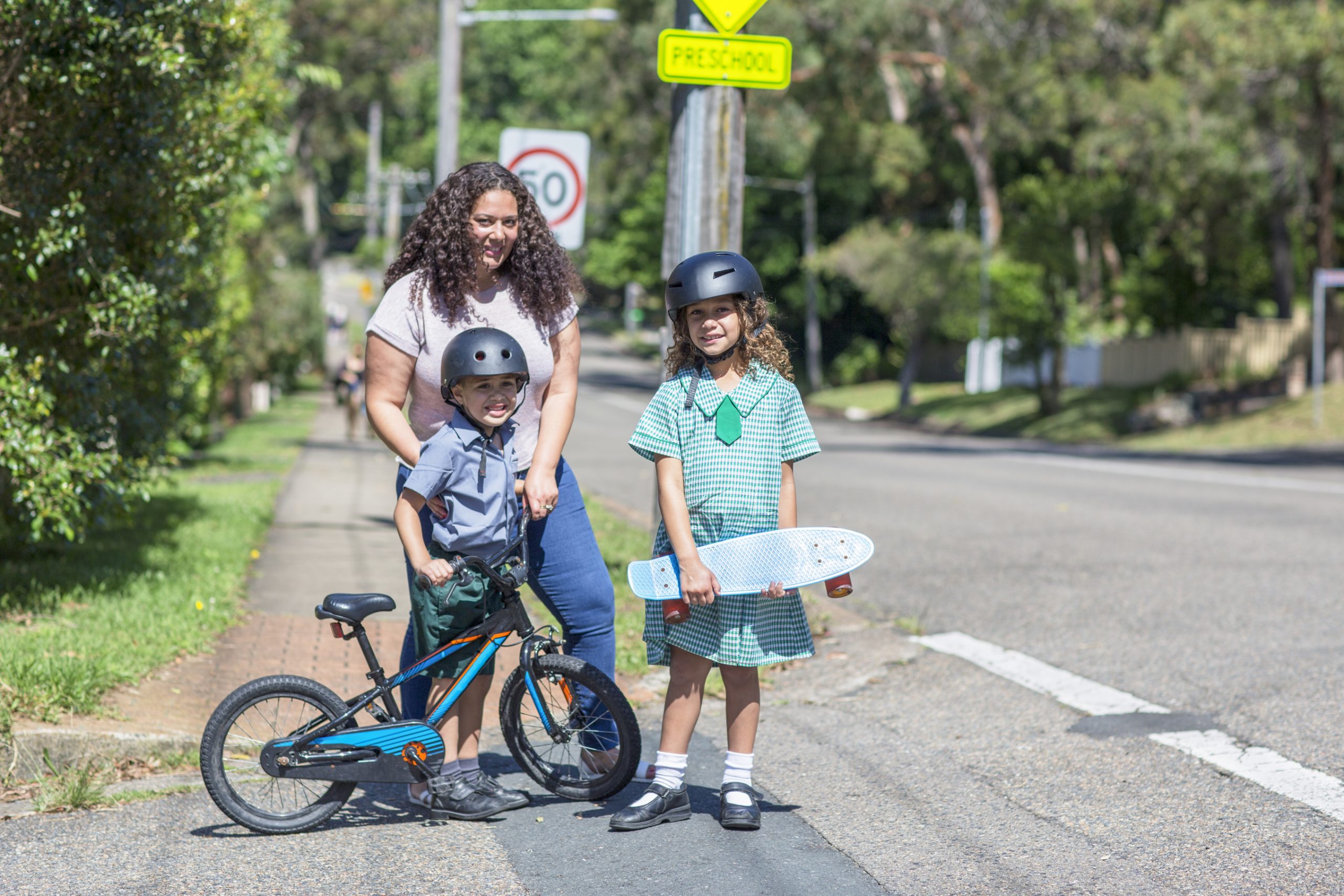 Mum and children crossing the road to school Woman with long dark hair standing on a road, with two children - a young boy aged about 6 years old on push bike and wearing a helmut, a young girl aged 11 years old wearing her school uniform, a helmut and carrying a skateboard