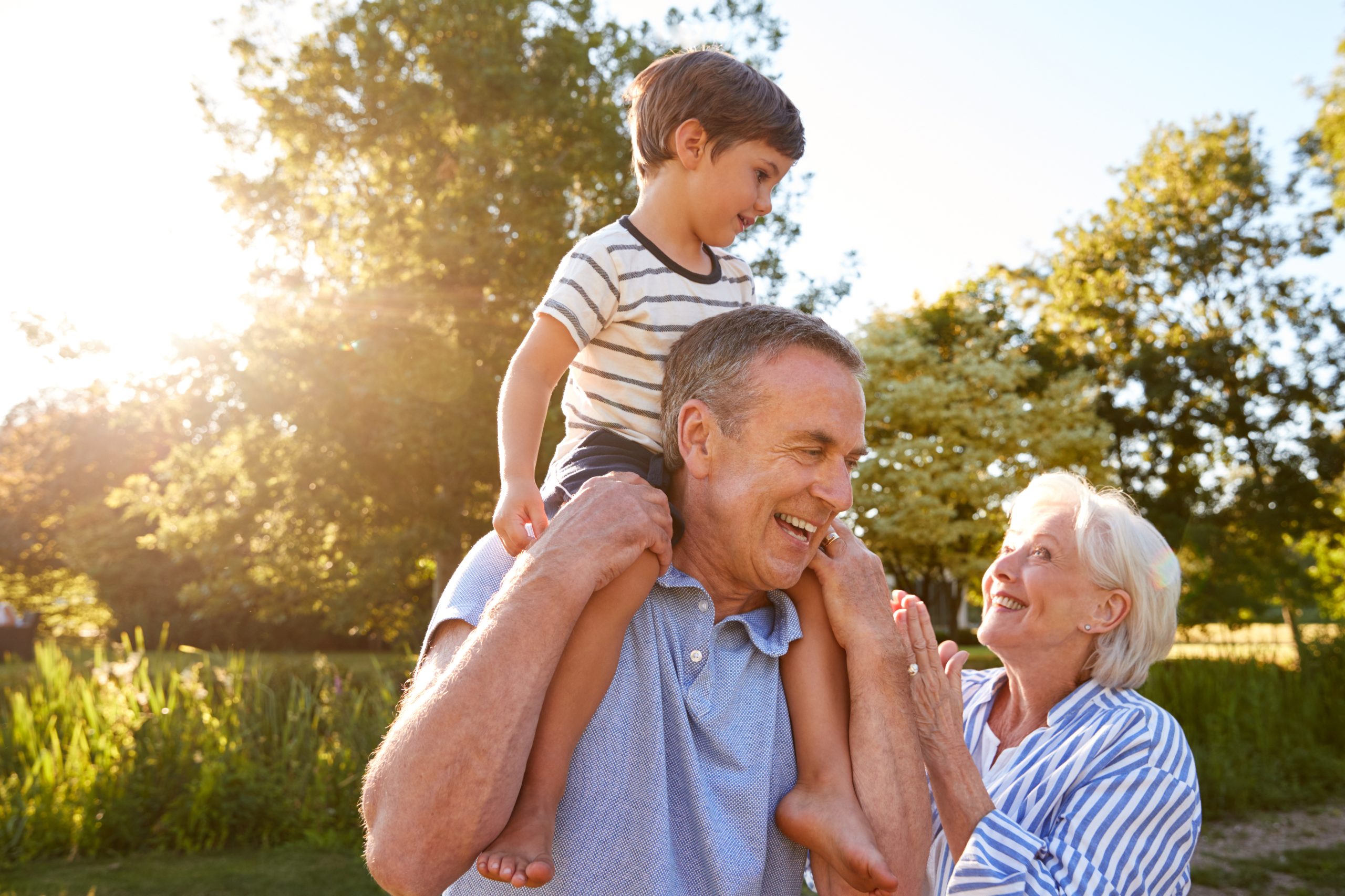Grandparents,Giving,Grandson,Ride,On,Shoulders,In,Summer,Park,Against